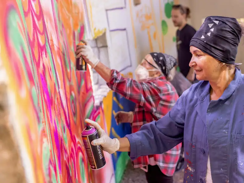 Zwei ältere Frauen - eine im karierten Hemd, die andere in blauer Arbeitsjacke - besprayen eine bunte Wand mit Gfaffiti. Sie tragen beide Tücher auf dem Kopf, eine trägt zusätzlich einen Mundschutz.