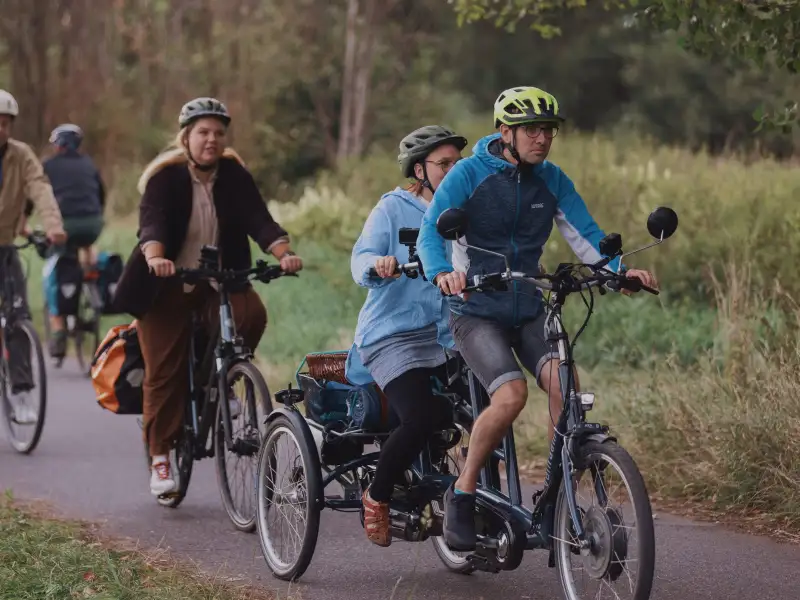 Ein Dreirad-Tandem mit zwei Personen fährt vor einer Radfahrerin auf einem Waldweg.