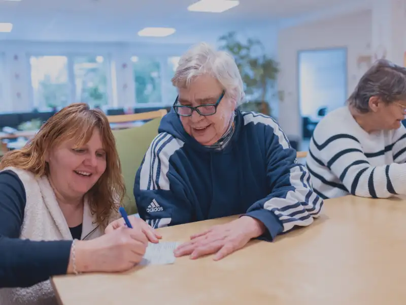 In einem Raum einer Begegnungsstätte: An einem Tisch sitzen mehrere älterer Menschen mit Behinderung. Eine Mitarbeiterin der Begegnungsstätte sitzt bei ihnen und verfasst mit den Teilnehmer*innen zusammen Postkarten.