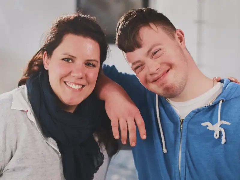Ein Mann mit Downsyndrom und eine Frau lächeln in die Kamera.