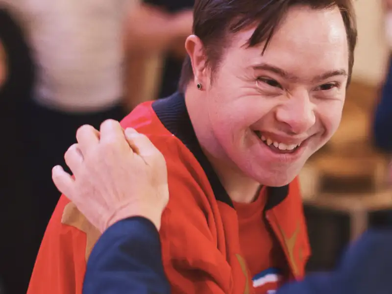 Ein junger Mann mit Downsyndrom steht lachend in einer Gruppe Leute