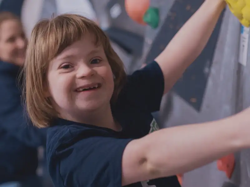 Ein Mädchen mit Downsyndrom klettert lächelnd an einer Boulderwand