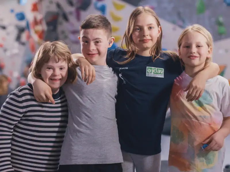 Vier Kinder mit und ohne Behinderung stehen nebeneinander vor einer Boulderwand