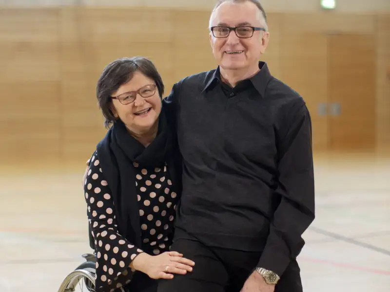 Ein älteres Paar in einer Sporthalle: Sie sitzt im Rollstuhl, er auf ihrem Schoß.