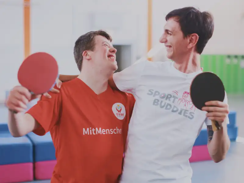 Ein junger Mann mit und einer ohne Downsyndrom stehen lachend Arm in Arm nebeneinander. Beide tragen Tischtennisschläger.