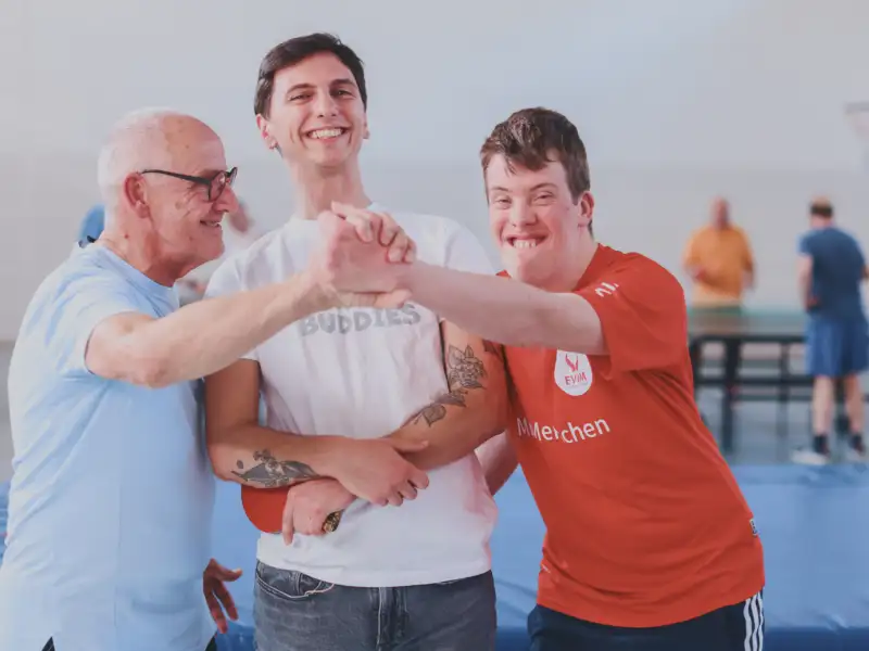 Drei Männer stehend lachend nebeneinander in einer Sporthalle, einer davon mit Downsyndrom