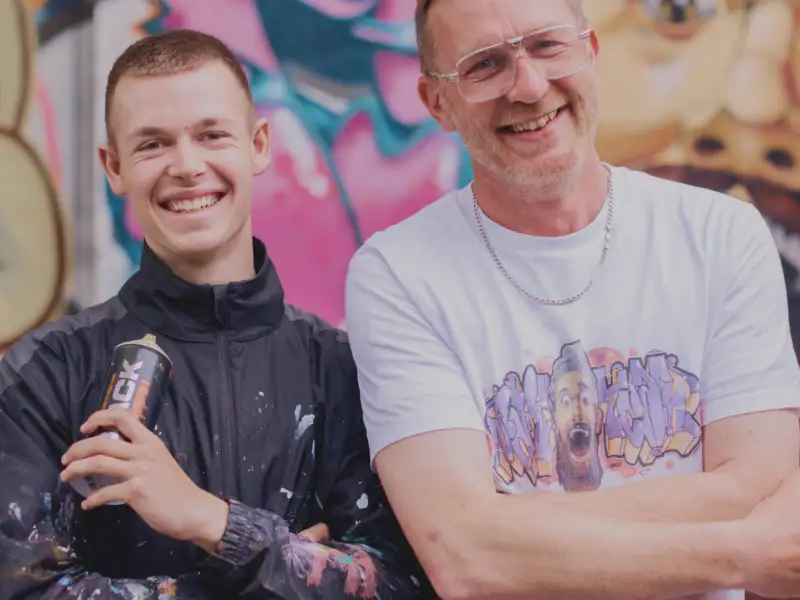 Ein Jugendlicher mit Spraydose und ein Mann stehen lächelnd vor einer Graffiti-Wand