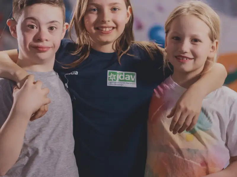 Zwei Mädchen und ein Junge mit Downsyndrom. Das Mädchen in der Mitte hat ihre Arme um die Schultern der anderen gelegt.