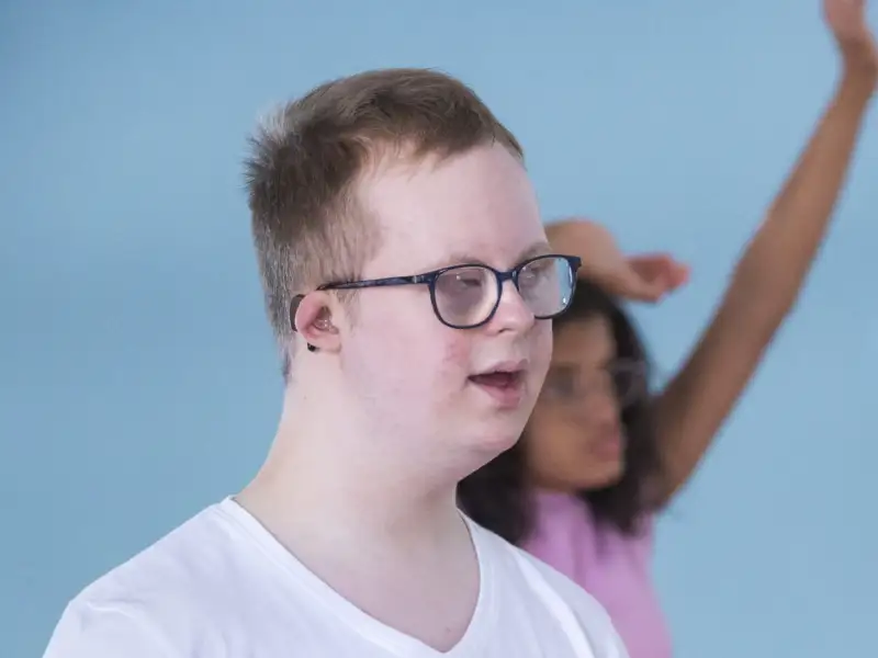 Ein junger Mann mit Downsyndrom, im Hintergrund tanzt eine junge Frau