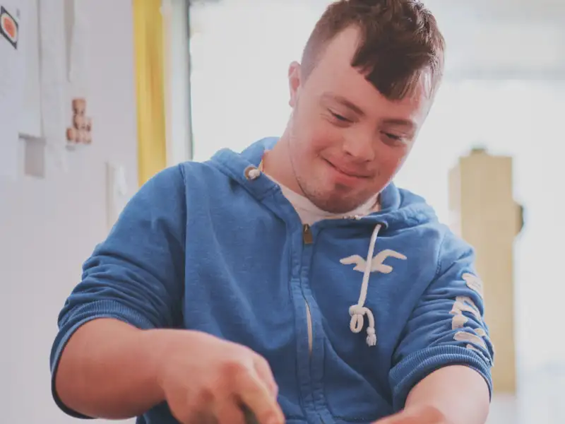 Ein junger Mann mit Downsyndrom im blauen Hoodie und modischer Kurzhaarfrisur schnippelt in einer Küche Zucchini.