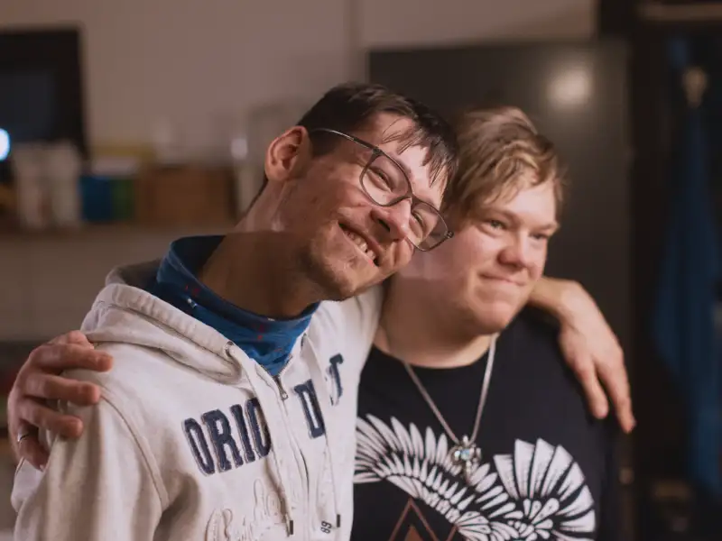 Zwei junge Männer mit geistiger Behinderung stehen lächelnd Arm in Arm nebeneinander.