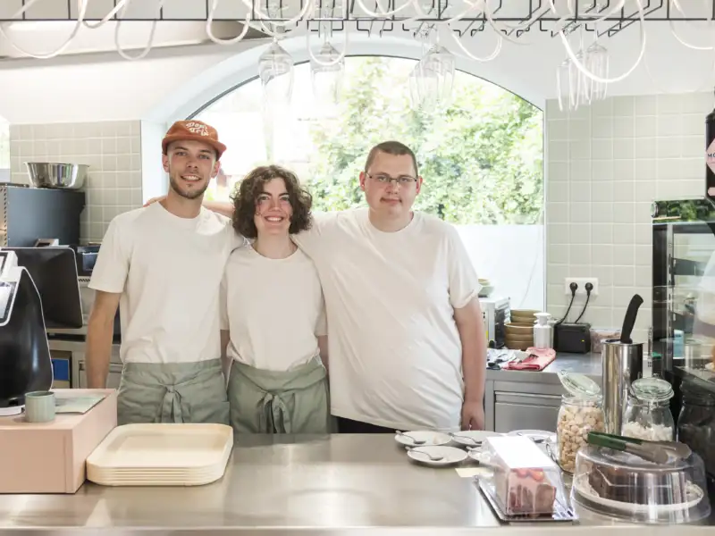 Eine Frau und zwei Männer stehend lächelnd hinter der Theke eines Cafés