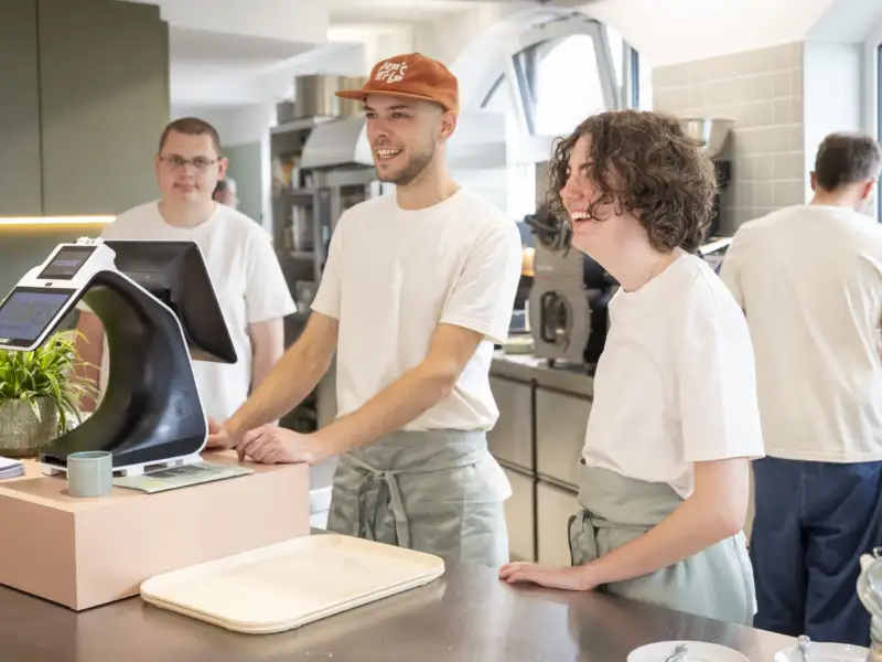 Drei Personen stehen hinter einer Theke mit Kasse eines Cafés