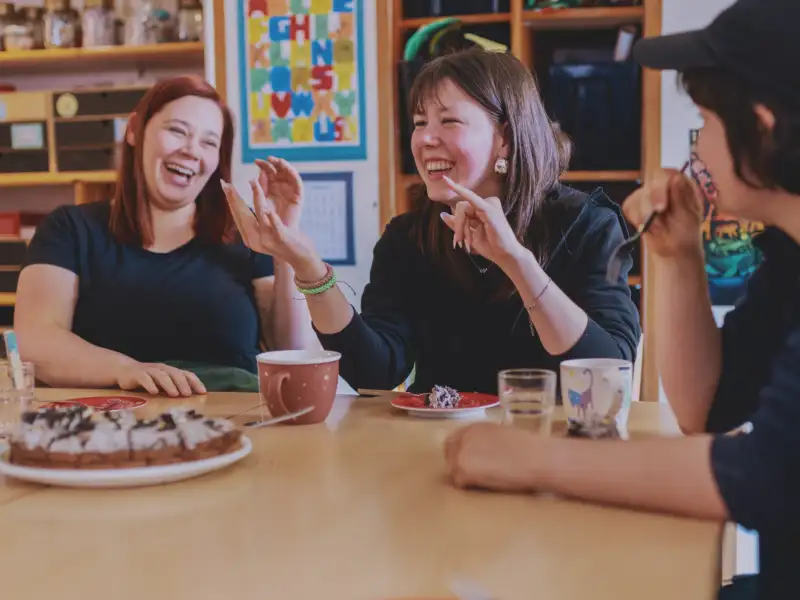 Drei junge Frauen sitzen bei Kaffee und Kuchen an einem Tisch und lachen miteinander.