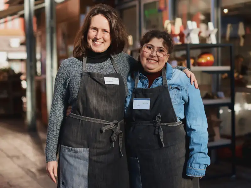 Zwei Frauen stehen vor einem Marktstand mit Deko-Artikeln. Sie tragen beide schwarze Schürzen und haben sich freundschaftlich die Arme um die Schultern gelegt. Beide lächeln in die Kamera.
