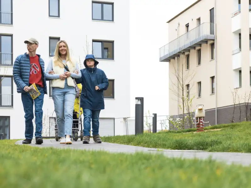 Eine Frau und ein Mann haben sich bei einer Frau in ihrer Mitte untergehakt. Die drei gehen über einen Weg zwischen Wohnhäusern.