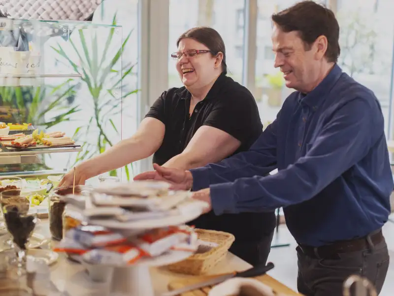 Eine Frau und Rudi Cerne stellen Schüsseln mit Frühstücksutensilien auf eine Theke.