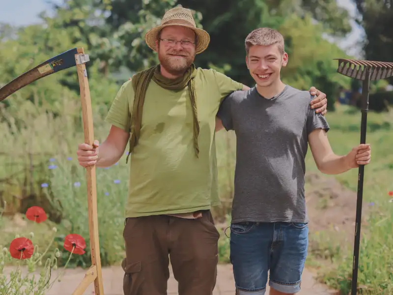 Zwei Männer stehen Arm in Arm nebeneinander mit Gartenwerkzeugen