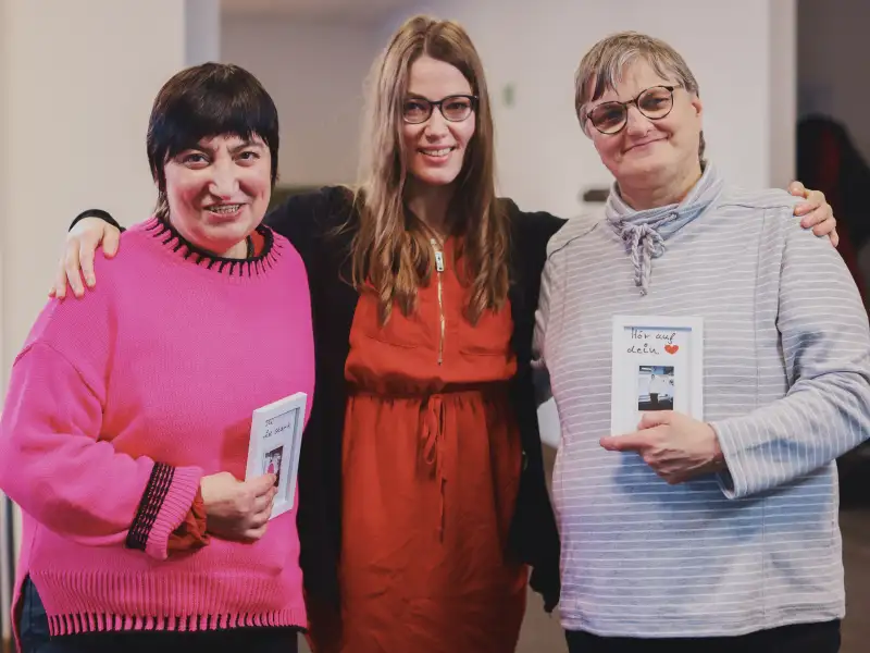 Drei Frauen stehen nebeneinander. Sie halten einen kleinen weißen Bilderrahmen in den Händen mit einem Foto von sich selbst und dem Satz "Hör auf dein Herz".