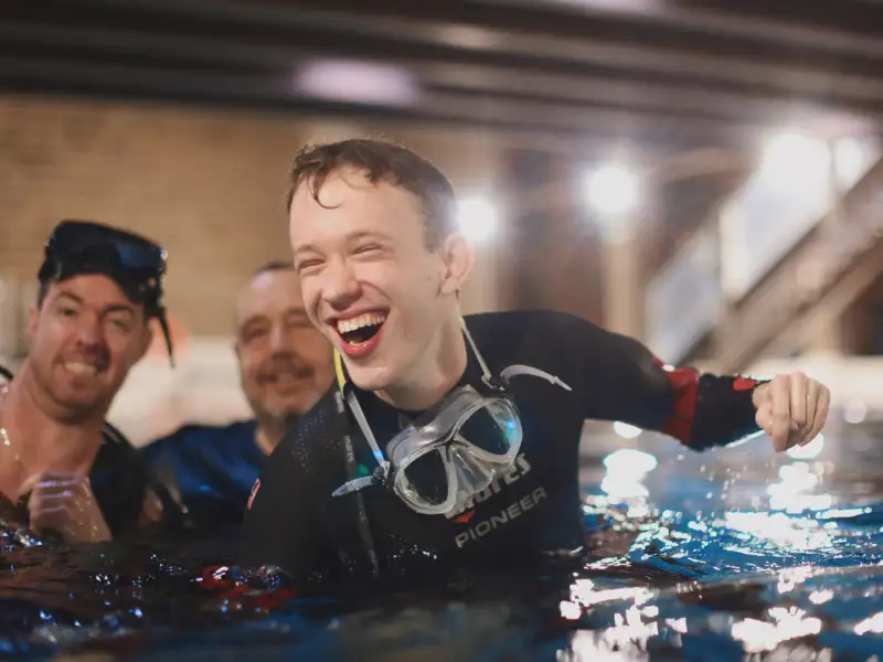 Drei Männer in Tauchkleidung schwimmen in einem Wasserbecken. Sie lachen.