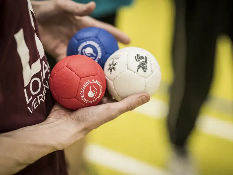 Ein Mann hält drei Boccia Kugeln in seiner rechten Hand. Die Kugeln haben die Farben rot, blau und weiß.