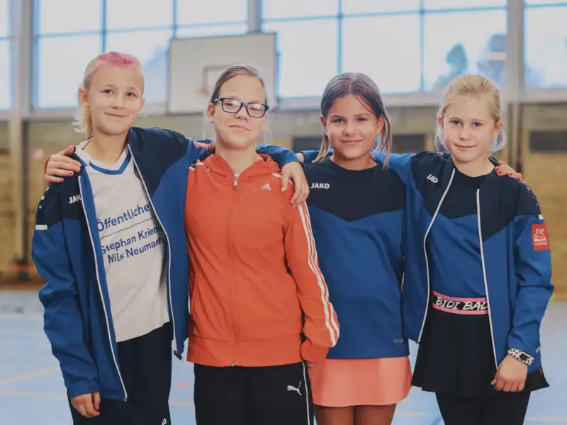 Vier Mädchen stehen nebeneinander. Sie haben die Arme um die Schultern gelegt und lachen.