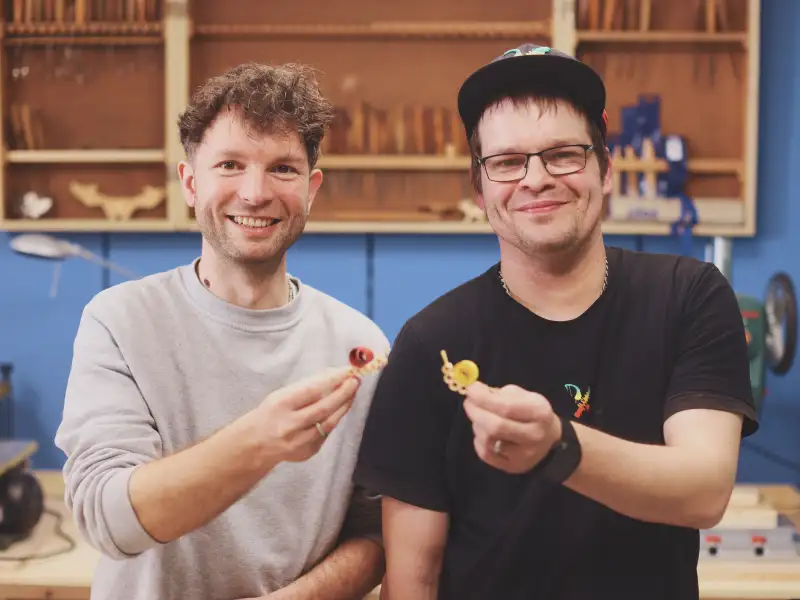 Gewinner Robert Berthold und ein junger Mann zeigen lächelnd ihre aus Holzspänen selbst gefertigten Schnecke vor.