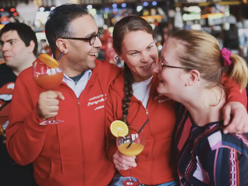Ein Mann und zwei Frauen stehen zusammen und lachen. Sie haben jeder einen Cocktail in der Hand.