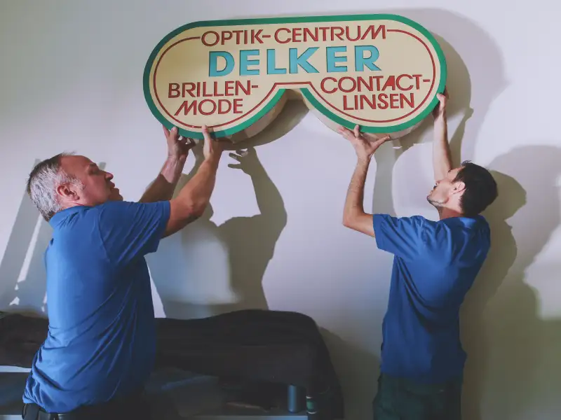 Zwei Männer montieren ein Schild an einer Wand. Auf dem Schild steht Optik-Centrum Delker.