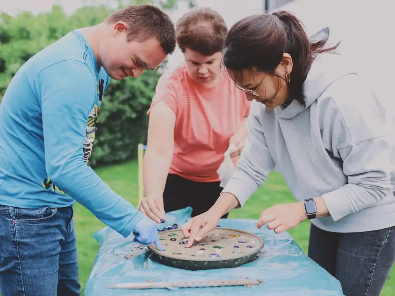 Zwei Frauen und ein junger Mann mit Down-Syndrom drücken bunte Steine eine Schale mit Sand.