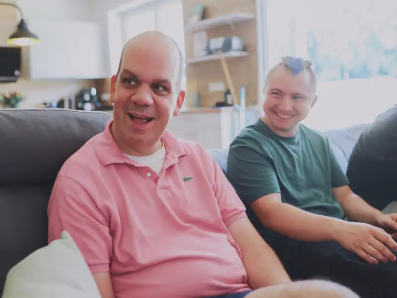 Zwei Männer sitzen auf einem grauen Sofa und lachen.