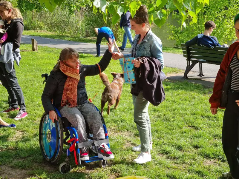 Zwei junge Frauen sprechen miteinander auf einer Wiese in einem Park. Eine von ihnen sitzt im Rollstuhl. Im Hintergrund läuft ein Hund.