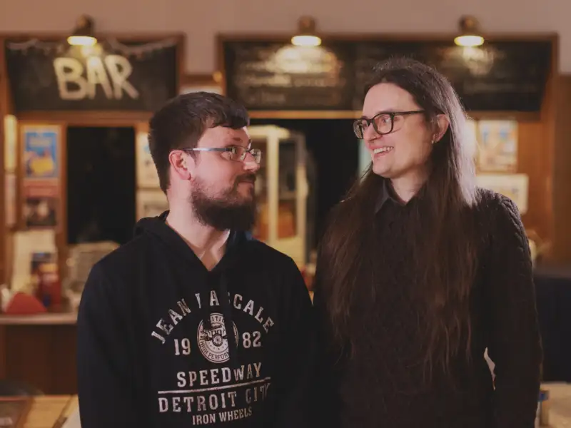 Zwei junge Männer stehen vor einer Bar und lächeln sich kumpelhaft an.