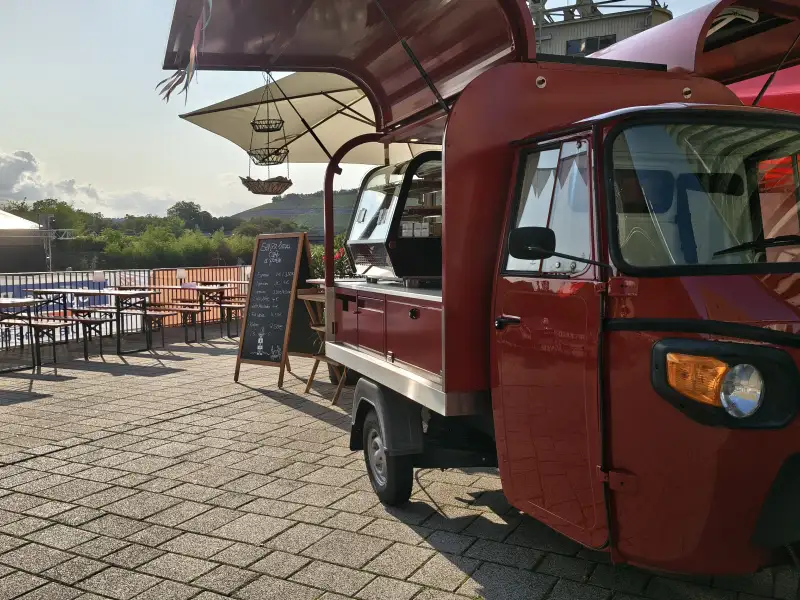 Ein Fahrzeug mit einer Kaffeebar parkt auf einer Terrasse. Im Hintergrund stehen Tische und Bänke.