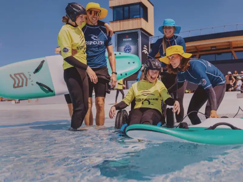 Eine Frau schiebt ein Surfbrett zu einer anderen jungen Frau, die sich im Rollstuhl nähert. Drei weitere Personen stehen dabei.