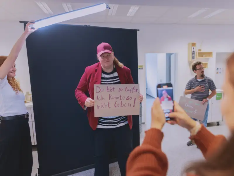 Ein großer Mann hält ein Schild vor seinen Körper auf dem steht: "Du bist so tapfer. Ich könnte so nicht leben!" und wird dabei gefilmt