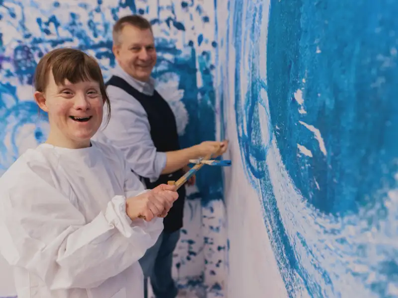 Eine junge Frau in weißem Kittel steht vor einer Wand, auf die sie mit einem Pinsel blaue Kreise malt. Im Hintergrund steht ein älterer Mann.