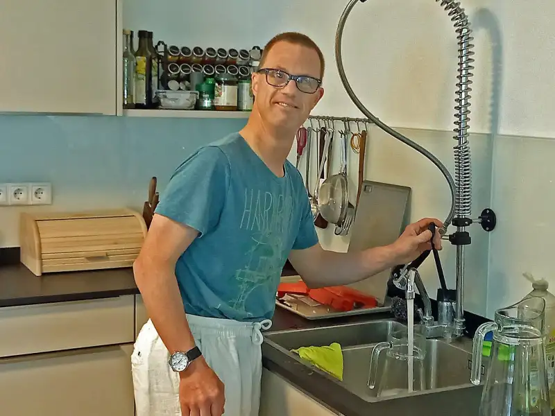 Ein Mann steht an der Spüle einer Küche. Er trägt ein T-Shirt und eine Brille.