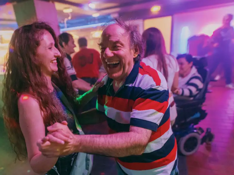 Auf der Tanzfläche in der Disco. Im Vordergrund tanzt ein älterer Mann mit einer jüngeren Frau. Im Hintergrund sind weitere Personen, darunter eine Frau im Rollstuhl zu sehen.