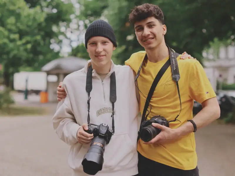 Zwei junge Männer mit Fotokameras auf einer Straße. Einer hat den Arm um den anderen gelegt.
