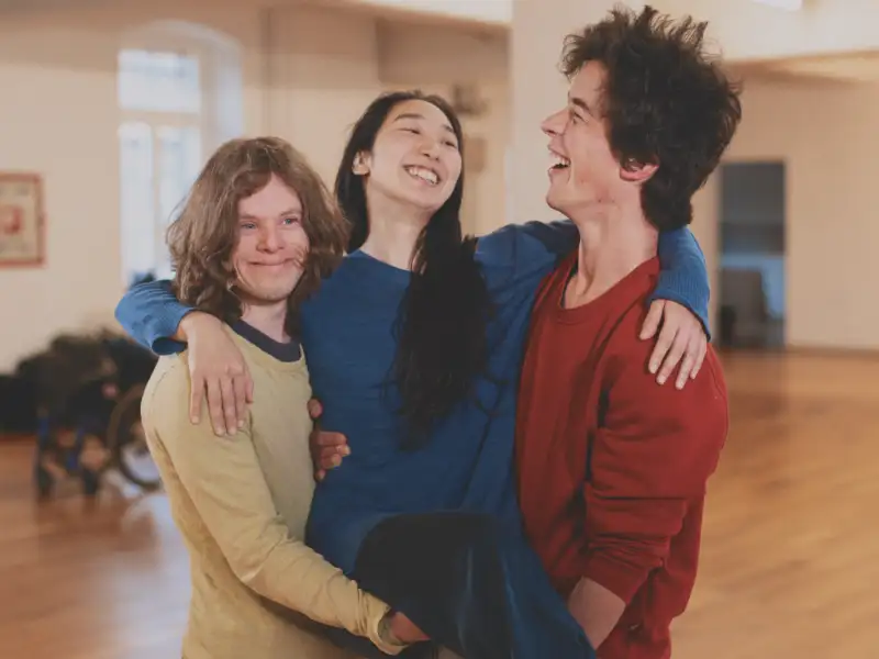Drei Personen stehend lachend Arm in Arm in einem Tanzstudio