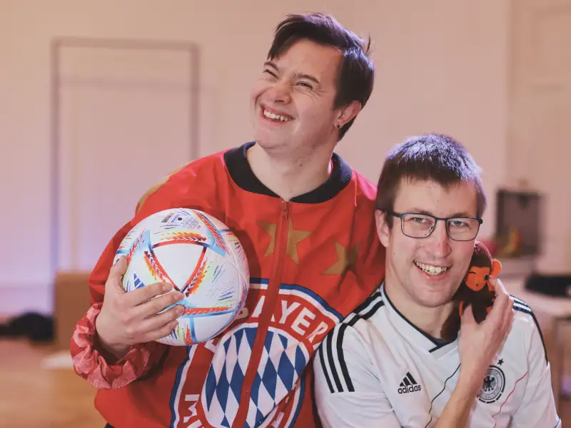 Zwei junge Männer, einer mit Downsnyndrom, mit Fußball-T-Shirt und einem Ball in einem Raum.