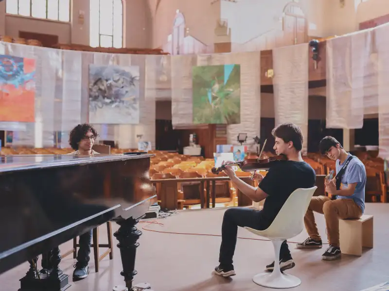 Drei junge Männer sitzen auf einem Podest in einer Kirche und spielen ihre Instrumente: Klavier, Geige und E-Gitarre.