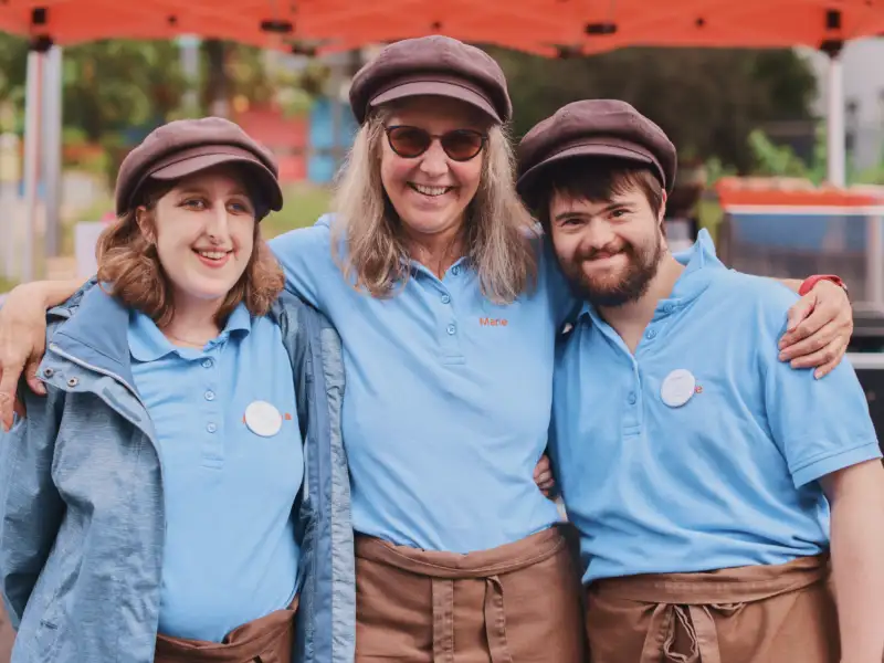 Zwei Frauen und en Mann stehen nebeneinander. Sie tragen braune Kappen und blaue T-Shirts. Im Hintergrund ist eine mobile Kaffeestation zu sehen.