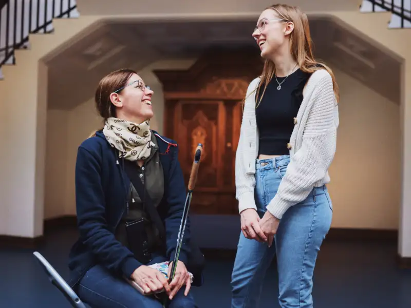 Zwei junge Frauen stehen im Foyer des Museums. Eine Frau hält einen Gehstock in der Hand.