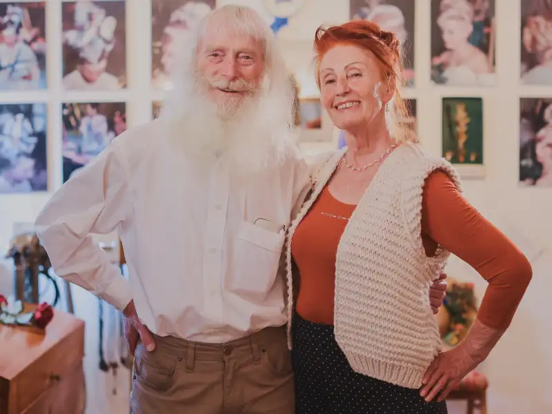 Ein Mann mit langem weißen Bart und eine Frau stehen nebeneinander und lachen in die Kamera.