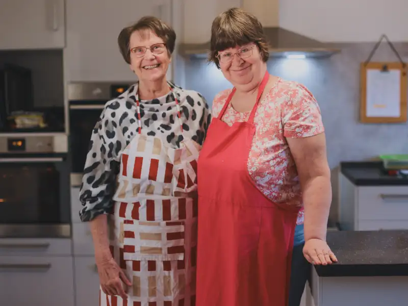 Zwei Frauen stehen nebeneinander in einer Küche. Sie lachen in die Kamera.