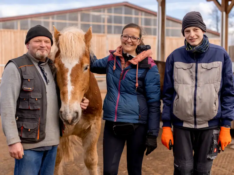 Zwei Männer, eine Frau und ein Pony vor einer Reitanlage.