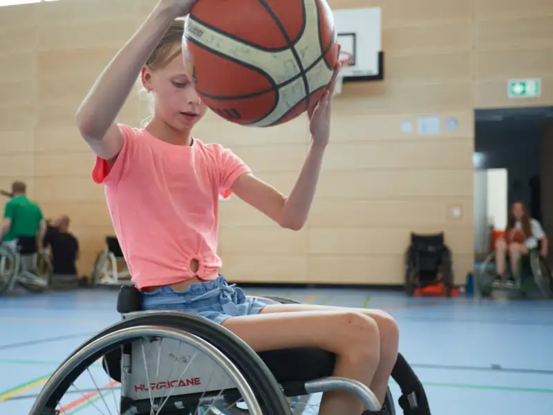 Ein Mädchen im Rollstuhl hält einen Basketball hoch.