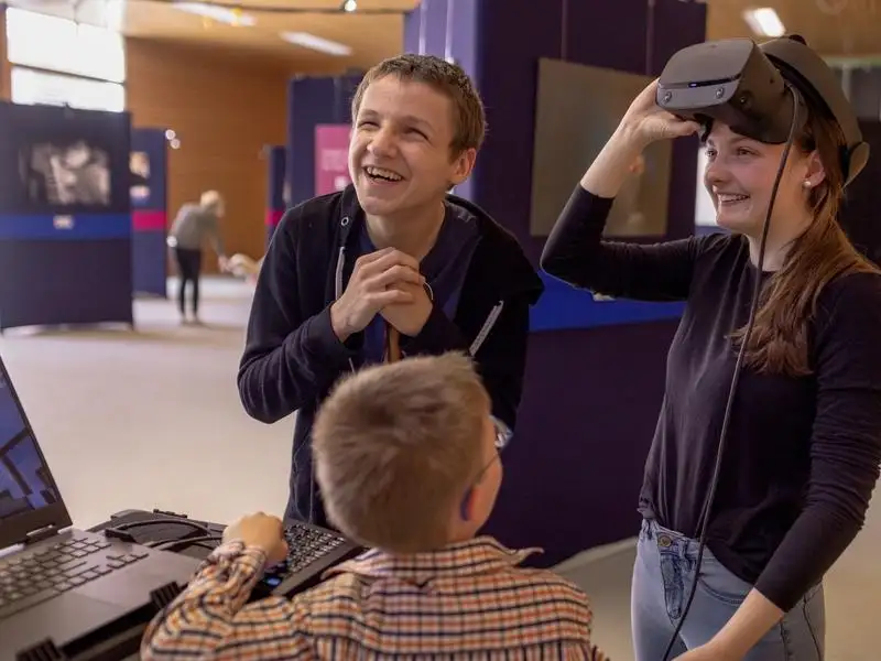 Drei Kinder und Jugendliche in einer Ausstellung, davon ein Mädchen mit VR Brille